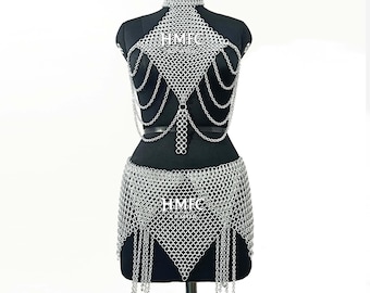 Könnte beinhalten: Ein silberfarbenes Kettenhemd-Set bestehend aus Oberteil und Rock mit hohem Kragen, komplexen ineinandergreifenden Metallringen und hängenden Kettenfransen. Der Text HMFC ARMOR ist auf dem Oberteil und dem Rock des Kleidungsstücks sichtbar.
