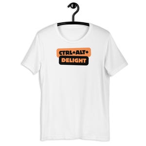 Puede incluir: Camiseta blanca con el texto "CTRL+ALT+DELIGHT" en naranja y negro. El texto está dispuesto en dos líneas horizontales, con "CTRL+ALT+" en la parte superior y "DELIGHT" debajo. La camiseta está colgada de una percha negra.