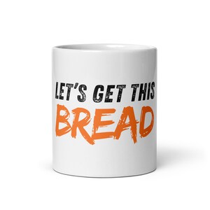 Può includere: Tazza in ceramica bianca con la scritta "LET'S GET THIS BREAD" in lettere nere e arancioni. La tazza è cilindrica e ha una finitura lucida. Il testo è in uno stile a pennellate audaci.