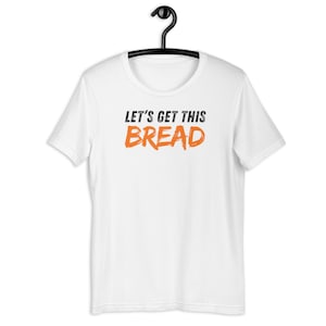 Può includere: T-shirt bianco con la scritta "LET'S GET THIS BREAD" stampata in nero e arancione. La maglietta è appesa a una gruccia nera. Il testo è in grassetto, stile pennellata.