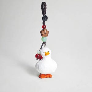 May include: A white duck-shaped keychain with orange feet and a yellow beak. The keychain has a brown cord with decorative beads in red, green, and brown. The duck is carved with visible wood grain.