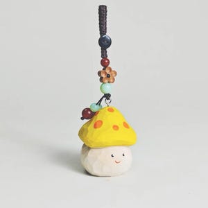 May include: A whimsical wooden mushroom charm with a yellow cap and orange spots, a smiling face, and a beaded cord. The charm features a brown cord, a dark blue button, wooden flower, and various colored beads.