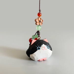 May include: A carved wooden cat charm with black and white fur, pink ears and paws, and blue eyes. The charm is suspended by a brown cord with decorative beads and a wooden flower.