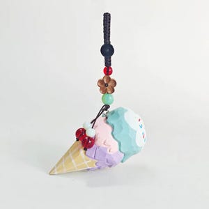May include: A colorful ice cream cone charm with a light yellow waffle cone, topped with scoops of pink, purple, and blue ice cream. The charm is adorned with red and white beads, a flower, and a black cord.