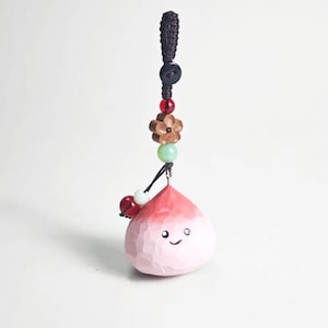 May include: A peach-shaped wooden charm with a smiling face, painted in shades of pink and red. The charm is suspended from a black cord with decorative beads, a wooden flower, and a black button. The charm is a unique accessory.