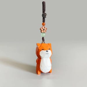 May include: A wooden fox figurine with an orange body and white chest. The fox is attached to a brown cord with a button, flower, and beads. The figurine is a keychain or charm.