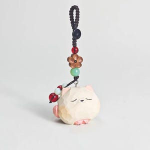May include: A carved wooden cat figurine with a serene expression, featuring pink accents on the ears and paws. The cat is suspended by a black cord adorned with beads in red, green, and brown, and a wooden flower. A unique, handcrafted cat charm.