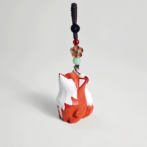 May include: A hand-carved wooden fox figurine with a red and white color scheme. The fox is suspended from a dark brown cord with decorative beads and a flower charm. The figurine is a unique decorative item.