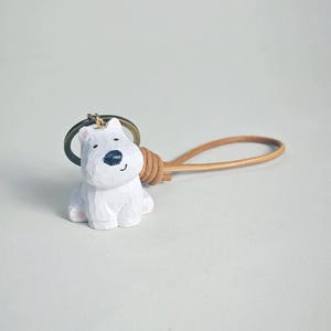 May include: A white, carved wooden polar bear keychain with a black nose and smiling mouth. The keychain has a brown leather strap and a metal ring. The bear's paws are painted pink.