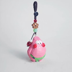 May include: A pink, carved wooden Patrick Star-inspired charm with a green swimsuit. The charm is suspended from a black cord with a button, beads, and a wooden flower. The charm has red ear muffs and a sad expression.