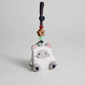 May include: A white cat-shaped charm with gray accents, including ears, paws, and a face. The charm is suspended from a dark brown braided cord, adorned with a button, beads, and a wooden flower. A cute, handmade accessory.