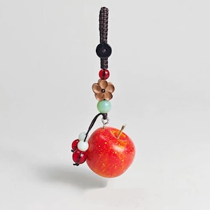 May include: A decorative apple-shaped charm with a brown woven loop, black button, red and green beads, and a bronze flower. A small cluster of red and white beads hangs below the apple. The apple is red with yellow spots.