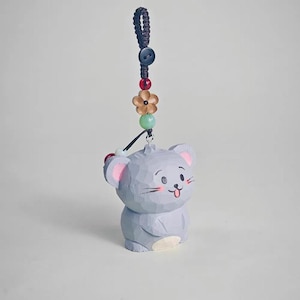 May include: A light gray, carved wooden mouse figurine with pink inner ears and a smiling face. The mouse is attached to a dark blue cord with a button, beads, and a flower charm. A cute, whimsical decorative item.