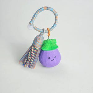 May include: A whimsical keychain featuring a purple eggplant charm with a smiling face and green leafy top. The keychain includes a colorful tassel and a braided loop in shades of blue, orange, and tan.