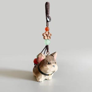 May include: A wooden dog figurine with a brown braided cord and decorative beads. The dog is light brown with a black collar and small red and green beads on its back. A wooden flower and orange bead are above the dog.