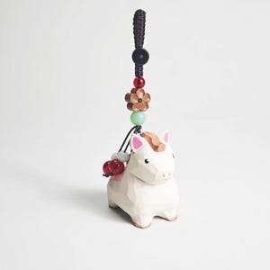 May include: A white wooden horse figurine with a brown mane and pink ears, attached to a decorative cord with beads and a flower. The horse is carved with geometric facets, giving it a unique, textured appearance. The cord is dark brown and black.