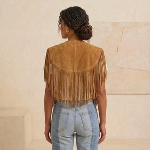 May include: A tan suede vest featuring long fringe detailing along the hem and back. The vest is worn over a plain white top and paired with light blue denim jeans. The garment has a curved back yoke and a sleeveless design with a soft texture.