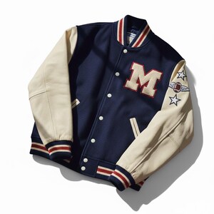 May include: Navy blue varsity jacket with cream-colored leather sleeves. Features a large letter "M" patch, white snap buttons, and striped cuffs and collar. Includes embroidered stars and a football graphic on the sleeve.