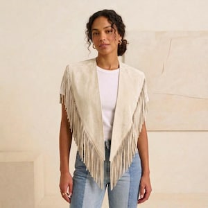 May include: A beige suede vest featuring a V-neckline and long fringe detailing along the front edges. The vest is worn over a plain white short-sleeved t-shirt and blue denim jeans, creating a layered look with a textured finish.
