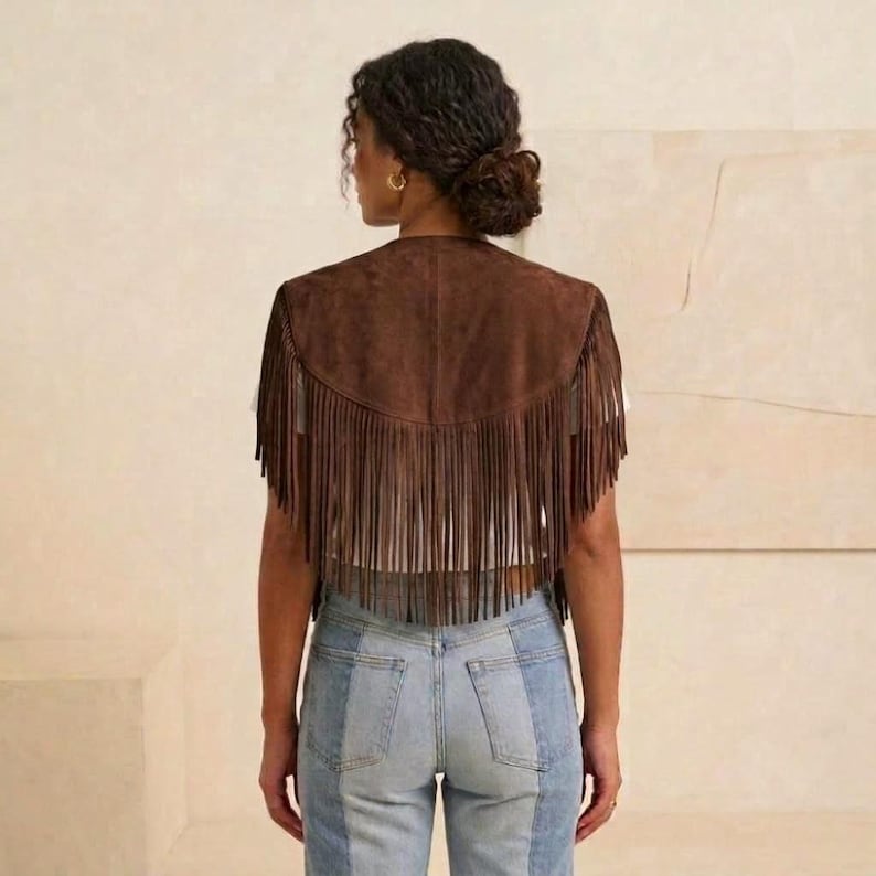May include: A dark brown suede vest featuring a V-shaped back yoke and long, vertical fringe detailing that extends from the hem. The vest is worn over a plain white top and paired with light blue denim jeans that have a visible vertical seam.