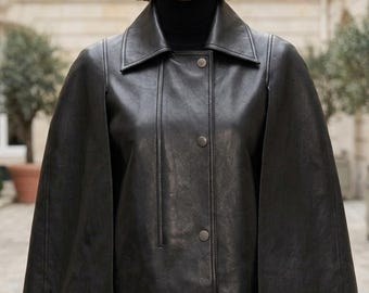 May include: A black leather cape jacket featuring a wide collar, a front button closure with three visible metallic snaps, and open arm slits. The garment is worn over a black turtleneck sweater, showcasing a smooth texture and structured silhouette.