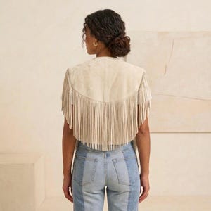 May include: A beige suede vest featuring a curved back yoke and long, dangling fringe trim along the hem and shoulders. The vest is worn over a plain white t-shirt and paired with light blue denim jeans that have a two-tone panel design.