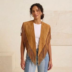 May include: A tan suede vest featuring a deep V-neckline and long fringe detailing along the front edges. The garment is sleeveless with a pointed hemline that extends down the torso, showcasing a soft texture and a structured, open-front design.
