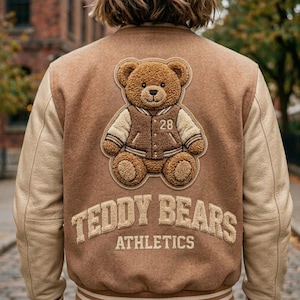 Op de afbeelding: Een beige varsity-jack met een grote, getextureerde chenille-patch van een teddybeer die een bijpassend jasje draagt met het nummer 28 op de borst. Onder de beer is de tekst TEDDY BEARS ATHLETICS geborduurd in grote, dikke, beige letters.