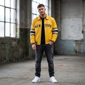 Può includere: Una giacca varsity in pelle gialla con la scritta NEW YORK in lettere nere sul petto, caratterizzata da polsini a righe bianche e nere, una t-shirt nera, jeans in denim grigio scuro consumati e scarpe da ginnastica bianche.