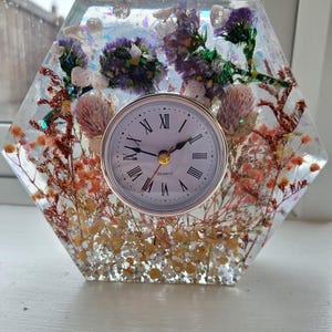 May include: A hexagonal clock with a clear resin body, showcasing dried flowers in shades of purple, pink, and orange. The clock face is white with black Roman numerals and gold hands. A unique decorative piece.