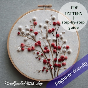 May include: A hand-stitched embroidery of a botanical design, set in a wooden hoop. The artwork depicts red berries and white fluffy details on brown branches, against a light fabric. Text includes "PDF PATTERN + step-by-step guide" and "beginner-friendly". The shop name "Pine Needle Stitch shop" is also visible.