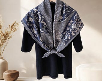 May include: A square scarf with a navy blue and gray floral pattern is draped over a black top. The scarf is knotted at the front and features intricate botanical and geometric designs. The scarf is made of a soft, flowing fabric.