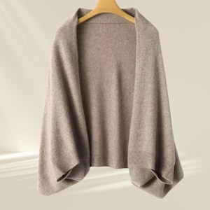 May include: A beige shawl or wrap, hanging on a wooden hanger. The garment features a draped design with wide sleeves and ribbed cuffs. The fabric looks soft and lightweight, ideal for layering. The colour is a neutral beige.