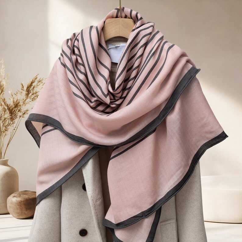 May include: A pink scarf with dark gray stripes and trim is draped over a coat. The scarf is made of a soft material and is hanging on a wooden hanger. The coat is a light beige color.