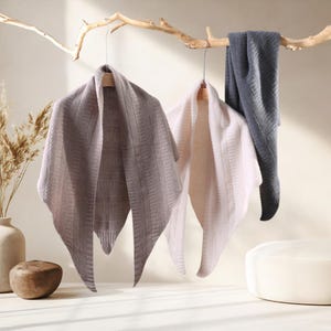 May include: Three triangular knit scarves hanging from a wooden branch. The scarves are in shades of light gray, soft pink, and dark charcoal gray. Each scarf features a textured, ribbed, and geometric knit pattern with pointed ends.
