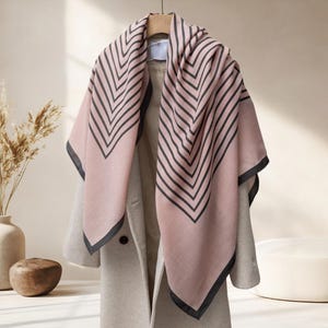 May include: A pink scarf with a black chevron pattern is draped over a light gray coat. The scarf has a dark gray border. The coat is hanging on a wooden hanger. The background is a neutral color.
