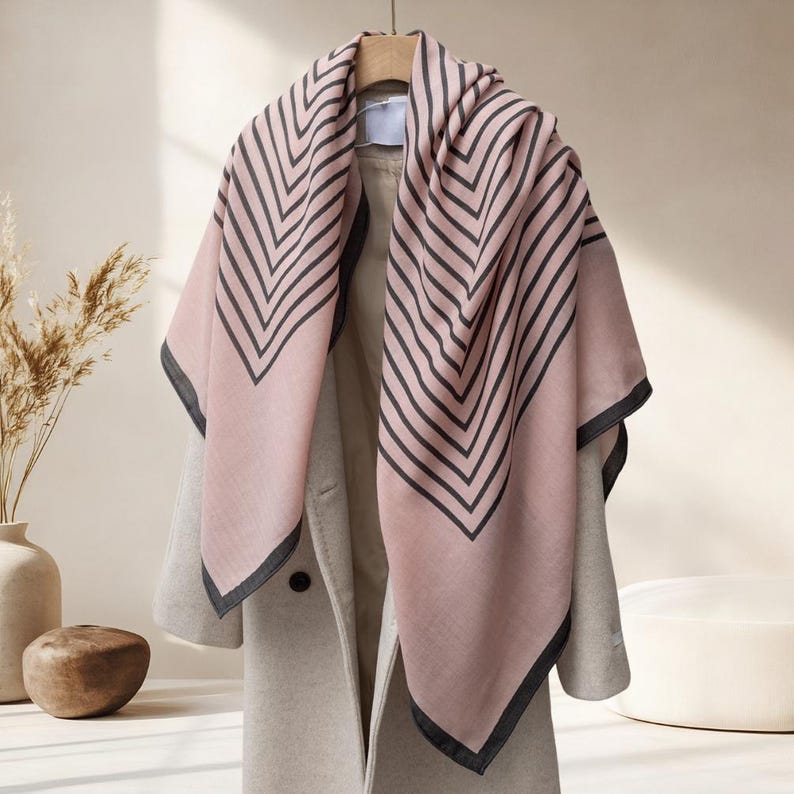 May include: A pink scarf with a black chevron pattern is draped over a light gray coat. The scarf has a dark gray border. The coat is hanging on a wooden hanger. The background is a neutral color.