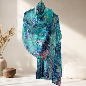 May include: A sheer scarf with a floral pattern in shades of teal, blue, and purple. The scarf is draped over a hanger, showcasing its flowing fabric and intricate design. The scarf's design features a watercolor-like effect.