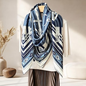 Op de afbeelding: Een vierkante sjaal met een marineblauw en wit patroon van riemen, gespen en paisley-motieven. De sjaal heeft een geometrische rand met kleine sterretjes en stippen, gedrapeerd over een lichtgekleurde top en een geplooide rok op een hanger.