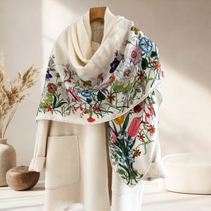 May include: A cream-colored scarf with a vibrant floral print featuring pink, blue, and yellow flowers, draped over a cream-colored coat. The scarf's design includes detailed botanical illustrations and a variety of colorful blooms.