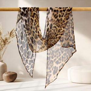 Pode incluir: Lenço transparente com estampa de leopardo. O tecido leve é drapeado sobre uma barra de madeira, mostrando manchas marrons, pretas e bege. O acessório de moda está pendurado, destacando o padrão animal em um fundo claro.