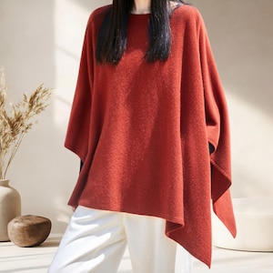 May include: A rust-colored, loose-fitting poncho with a wide, rounded neckline and an asymmetrical hemline. The garment features a soft, textured knit fabric that drapes over the shoulders and arms, paired with plain, light-colored trousers.