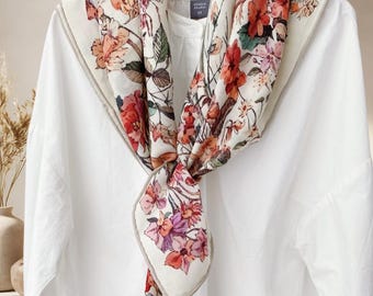 Puede incluir: Una camisa blanca con una bufanda floral sobre ella. La bufanda presenta un fondo crema con flores rojas, naranjas y moradas. La bufanda está anudada en la parte delantera, mostrando el colorido estampado floral.