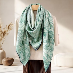 May include: A green and white patterned scarf with a leaf design, displayed on a wooden hanger. The scarf has a dark blue border and is draped over a cream-colored sweater. The overall aesthetic is elegant and suitable for various occasions.