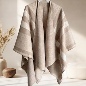 May include: A beige and cream colored wrap featuring a large plaid pattern with wide vertical and horizontal stripes. The garment has an open front design with draped fabric panels that hang down from a central point, creating a soft silhouette.