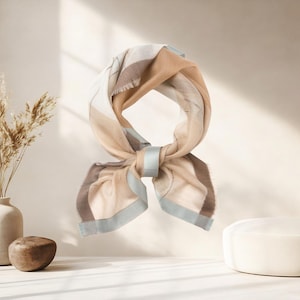 May include: A beige, brown, and white scarf with a light blue border and accents. The scarf is knotted at the bottom and appears to be made of a soft material. The scarf is displayed on a white surface.