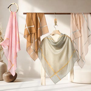 May include: A display of lightweight scarves in various colors, including pink, tan, and gray. Each scarf features metallic gold or silver stripes. The scarves are draped and hung, showcasing their flowing fabric and design.