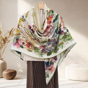 May include: A floral print scarf with a cream background. The scarf features a colorful botanical design with pink, purple, and blue flowers, green leaves, and a light green border. The scarf is draped over a hanger.