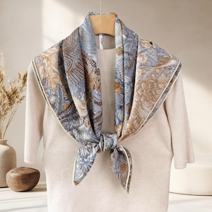 May include: A patterned silk scarf in blue, brown, and cream, knotted at the front. The scarf is draped over a beige sweater. The design features botanical elements. The scarf's measurements are in centimeters.