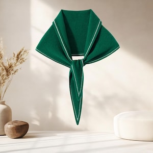 May include: A dark green triangular scarf featuring a wide collar-like fold at the top and a pointed tail at the bottom. The edges of the scarf are finished with a thin, white stitched border that runs along the perimeter of the fabric.